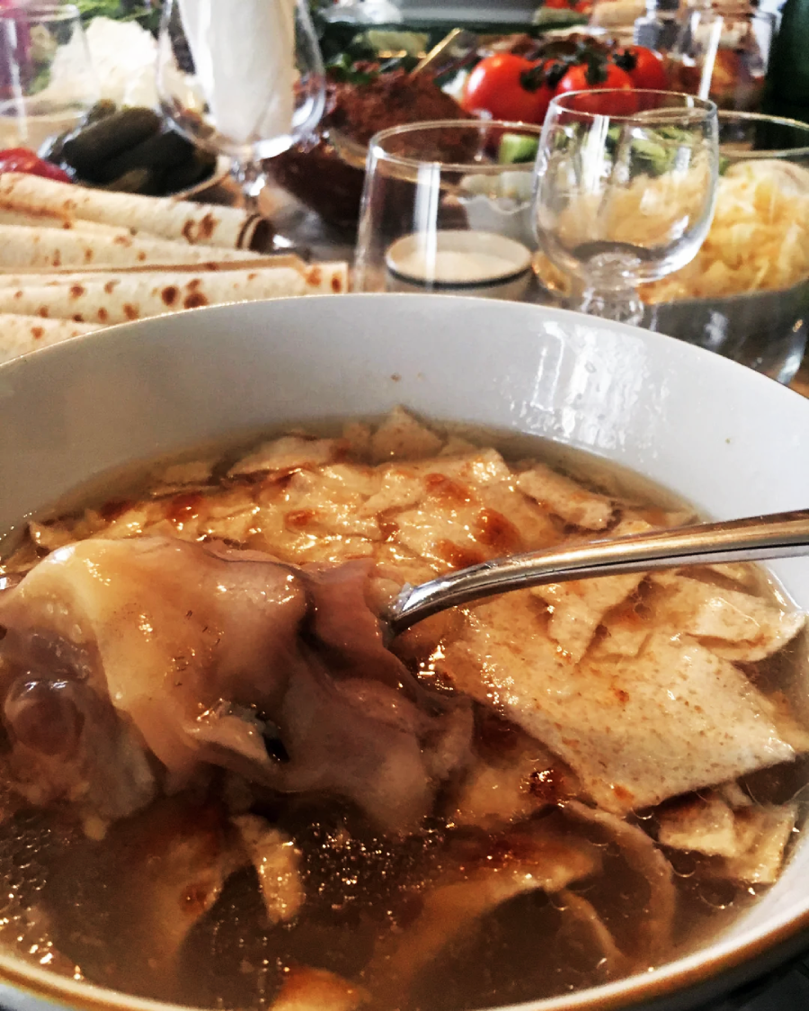 A bowl of soup with flatbread pieces and meat, served with a spoon. In the background, rolled flatbreads, vegetables, glassware, and side dishes are visible on a dining table.