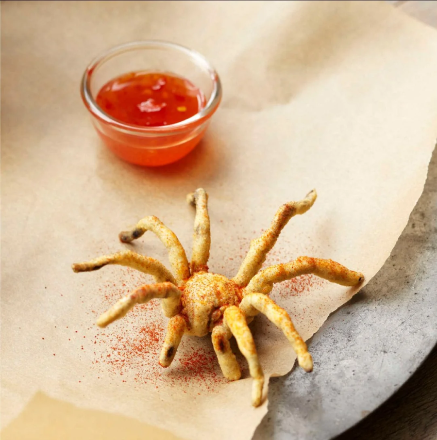 A deep-fried spider, dusted with seasoning, sits on parchment paper next to a small glass bowl of red dipping sauce.