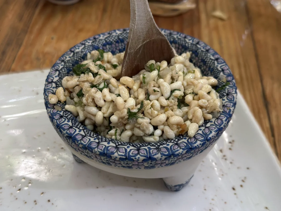 A decorative blue and white bowl filled with escamoles (ant larvae) mixed with herbs, with a wooden spoon resting inside, placed on a wooden table.