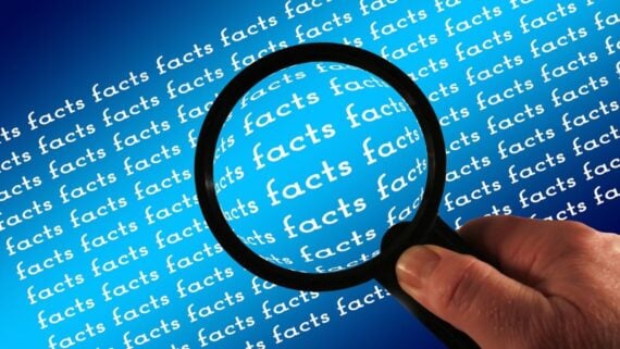 A hand holds a magnifying glass over the repeated word "facts" in white text on a blue background, enlarging the word "facts" in the center of the image.