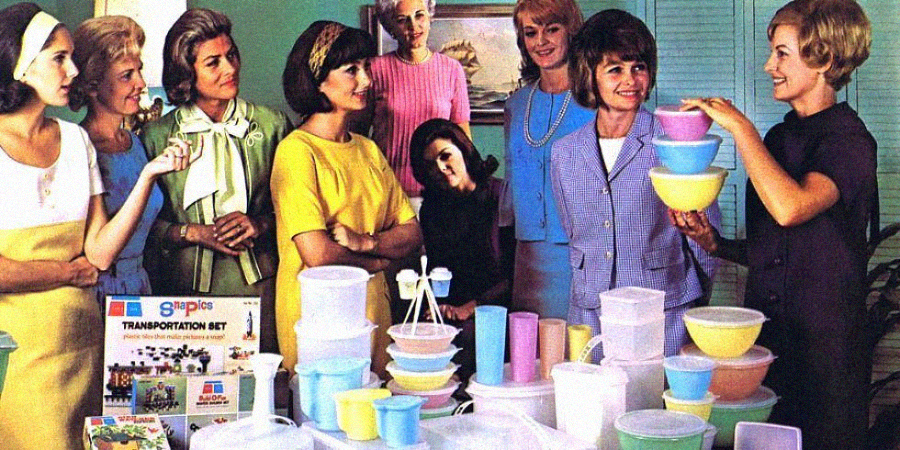 A group of women in 1960s-style clothing gather around a table full of colorful Tupperware containers, as one woman demonstrates the products and others watch and smile.