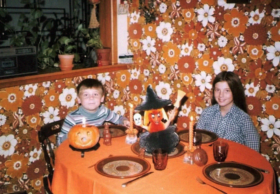 Two children sit at an orange table decorated for Halloween with pumpkins and festive figurines. The room has retro floral wallpaper, and plants are on the windowsill in the background.