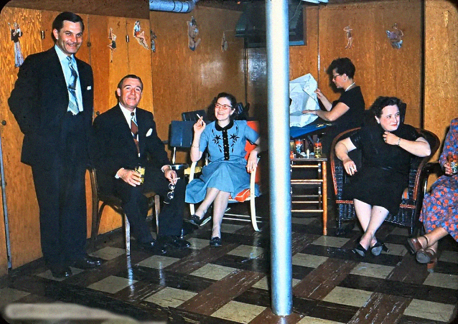 A vintage photo of five adults in formal and casual attire sitting and standing in a wood-paneled room with checkered flooring, chatting and relaxing. Decorations hang on the walls, and a metal support pole is in the center.