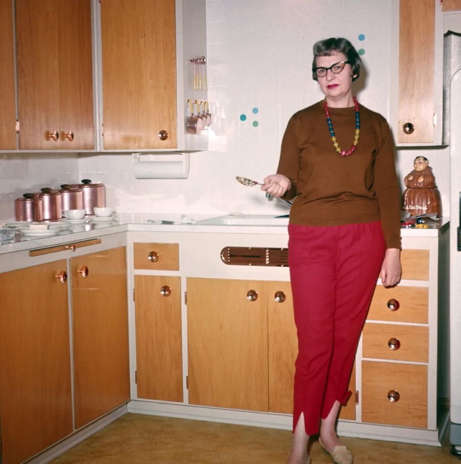 A woman in red pants and a brown sweater stands in a vintage kitchen with wooden cabinets, holding a spoon. She wears glasses and a colorful necklace. Copper canisters and a ceramic owl are visible on the countertop.