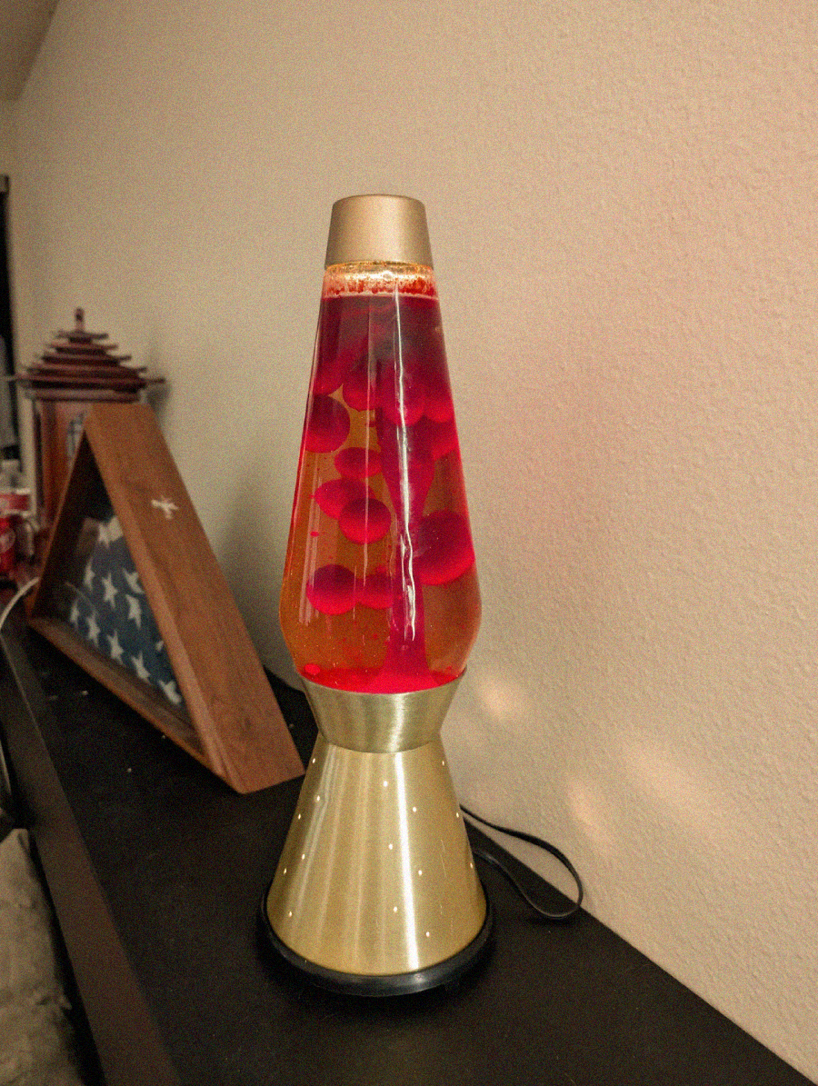 A gold-based lava lamp with red liquid and floating wax blobs sits on a black surface beside a folded American flag in a display case and a decorative jar against a beige wall.
