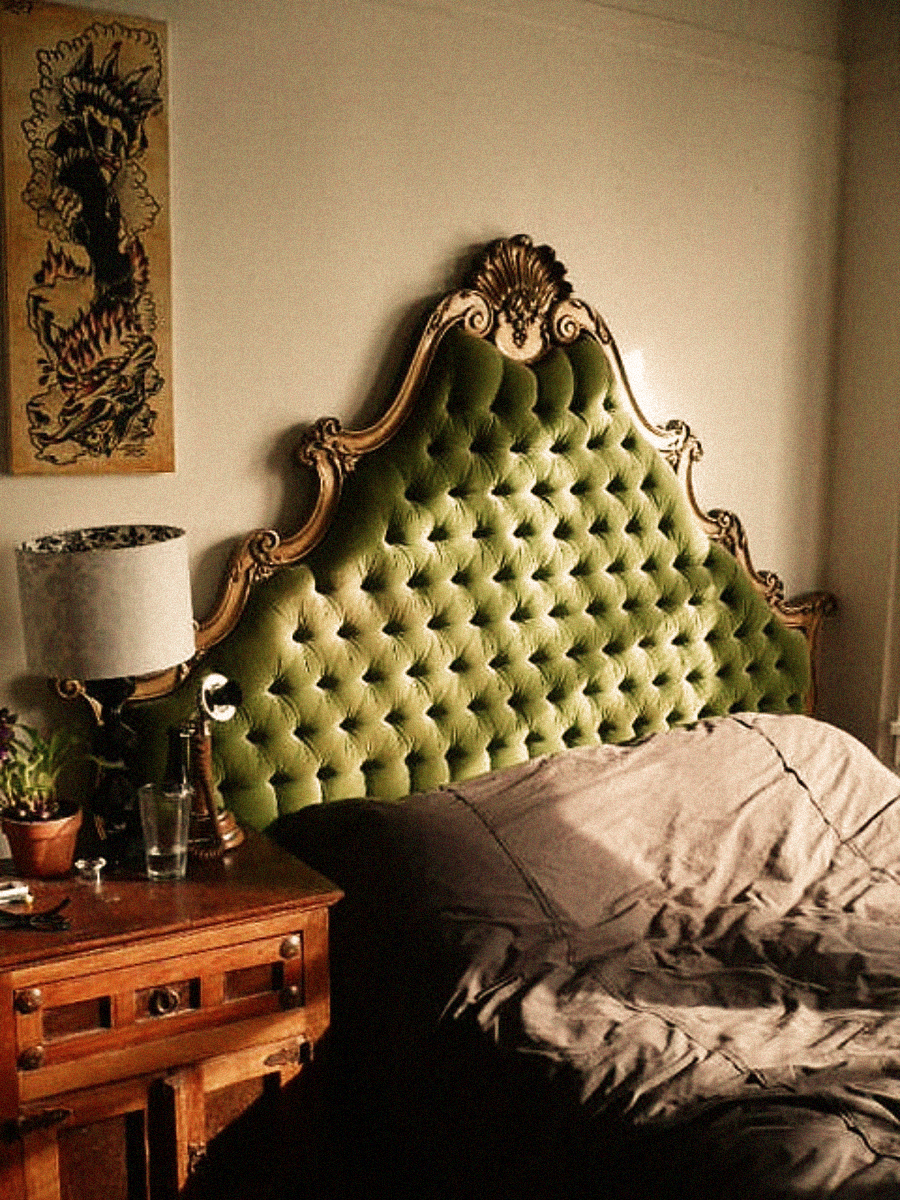 A vintage bedroom with an ornate, green tufted headboard, a wooden nightstand holding a lamp, glass, and plant, and an unmade bed with gray bedding. A framed artwork hangs on the wall above the nightstand.