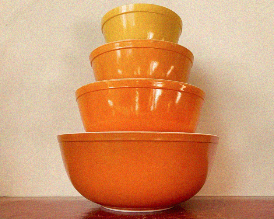 A set of four vintage mixing bowls, each a different size, stacked inside each other from largest to smallest. The bowls are in shades of orange and yellow and sit on a reddish surface against a plain white background.