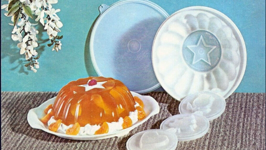 A molded gelatin dessert with fruit and whipped cream sits on a white plate. Behind it are two plastic molds—one upright, one open—plus four smaller lids. White flowers hang in the upper left corner against a blue background.