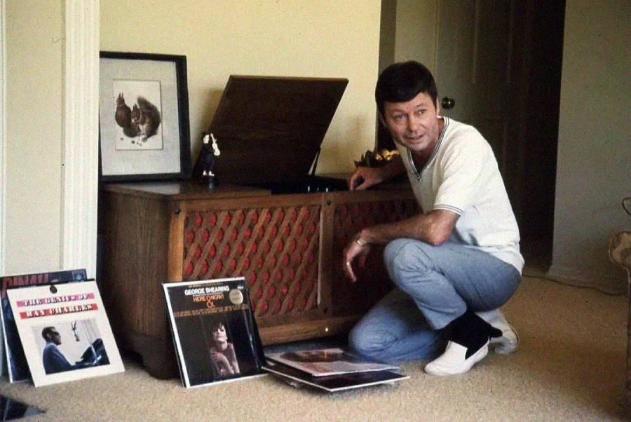 A man in a white shirt and light blue jeans kneels beside a vintage wooden record player, surrounded by vinyl records. There is framed artwork on the record player, and the setting appears to be a cozy room.