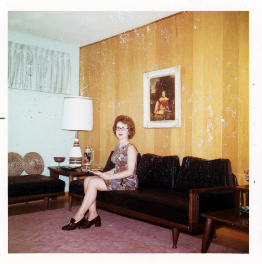 A woman with short curly hair and glasses sits on a dark sofa in a mid-century living room with a wood-paneled wall, lamp, framed artwork, and pink carpet. She is wearing a patterned dress and dark shoes.