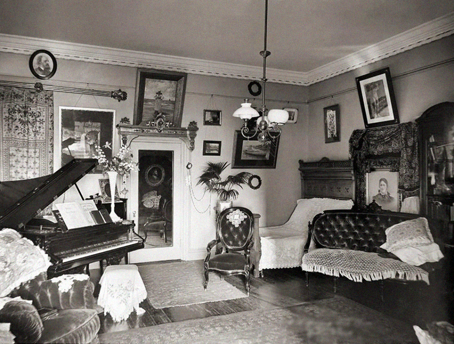 A vintage living room with ornate furniture, a grand piano, framed portraits on the walls, an elaborate mirror, a chandelier, and patterned rugs, creating an elegant, historical atmosphere.