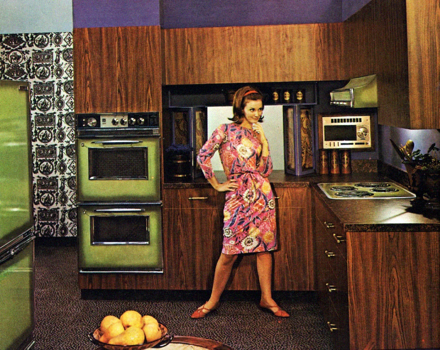 A woman in a colorful floral dress stands in a retro 1970s kitchen with wood cabinets, green appliances, and patterned tile walls. A bowl of oranges sits on a counter in the foreground.