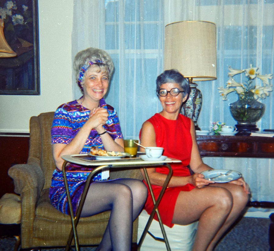 Two women sit indoors smiling, one in a colorful dress with a headband, the other in a red dress with glasses. They have food and drinks on trays in front of them, with a lamp and flowers on a table in the background.