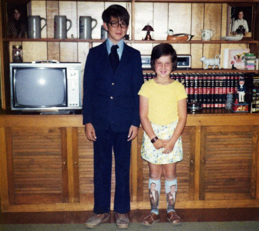 A boy in a blue suit and glasses stands next to a smiling girl in a yellow shirt, floral skirt, and novelty socks, in front of a wooden cabinet with a TV, books, and various decorative items.