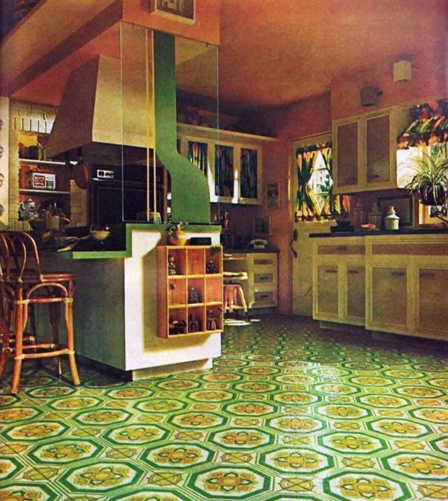 A retro-style kitchen with green and yellow geometric patterned floor tiles, white cabinets, a green vent hood, rattan bar stools, and bright sunlight filtering through floral curtains.