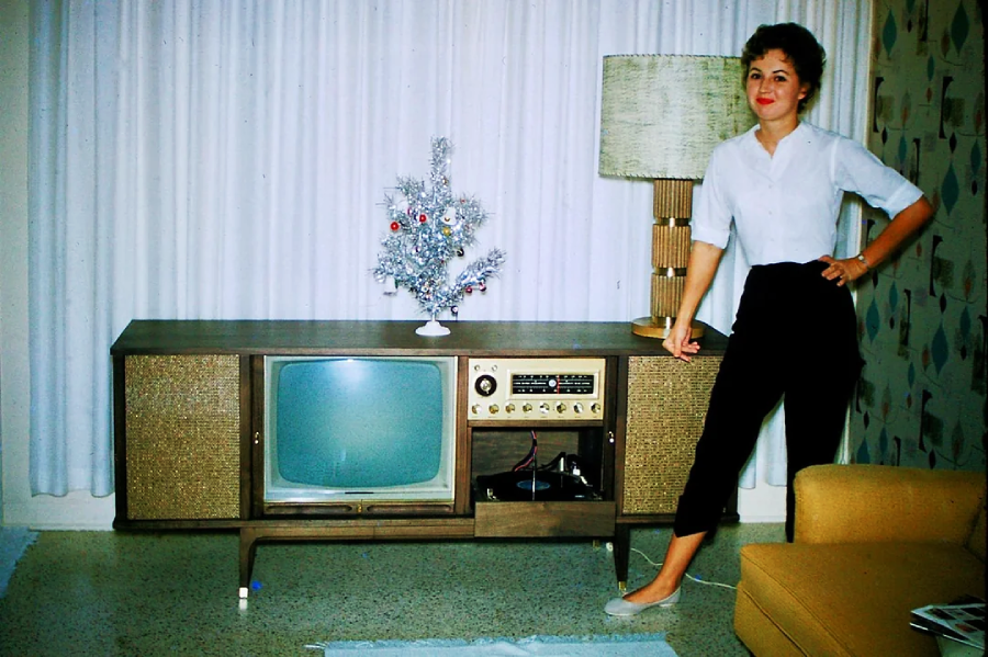 A woman in a white blouse and black pants stands beside a vintage console TV and radio, decorated with a small silver Christmas tree and a lamp, in a mid-century living room.