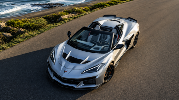 A silver convertible sports car with a black interior is parked on a coastal road, with the ocean and rocky shore visible in the background under clear skies.