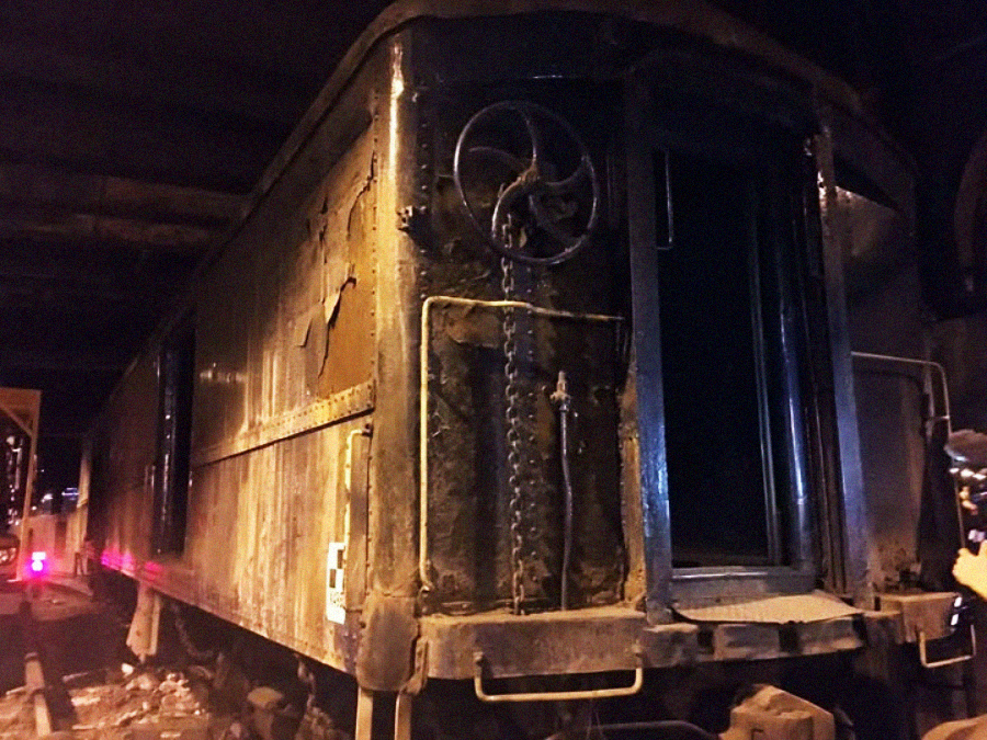 A close-up of an old, rusted train car at night, with a large metal wheel and chains on its rear. The scene is dimly lit, and a person with a camera is visible on the right edge of the image.