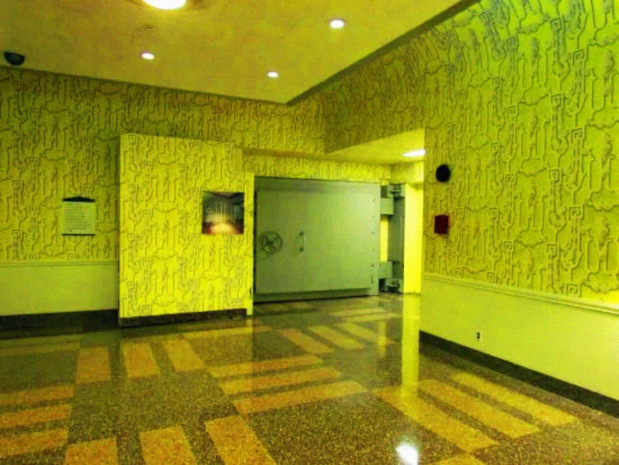 A brightly lit hallway with yellow wallpaper featuring abstract patterns, terrazzo floor tiles in brown and beige, and a large, silver double door in the background.