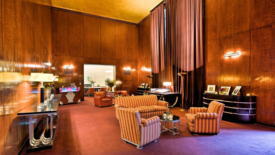 A spacious, elegant room with high ceilings, wood-paneled walls, striped upholstered sofas and chairs, modern lamps, framed photos on tables, and large floor-to-ceiling curtains. The lighting is warm and inviting.