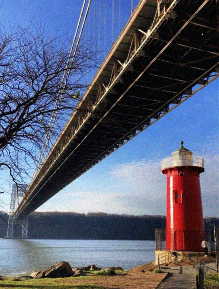 A bright red lighthouse stands on the riverbank beneath a large suspension bridge, with leafless tree branches on the left and blue sky above.