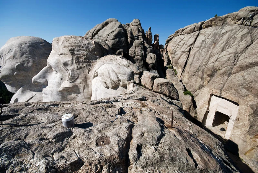 A behind-the-scenes view of Mount Rushmore shows the backs of the carved presidents’ heads and a rectangular doorway carved into the rock, surrounded by rugged stone formations under a clear blue sky.