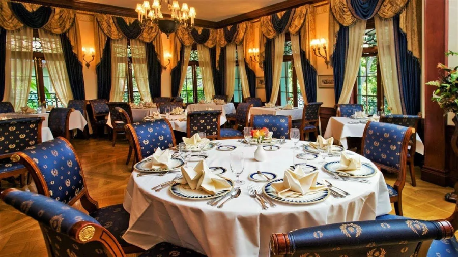 Elegant restaurant with blue and gold chairs, round tables set with white tablecloths, folded napkins, and fine china. Large windows with lavish drapes and chandeliers create a luxurious, classic atmosphere.
