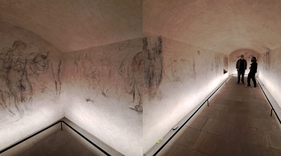 Two people stand in a softly lit underground corridor with stone floors and sketchy Renaissance-style drawings covering the curved plaster walls. The atmosphere is quiet and historical.