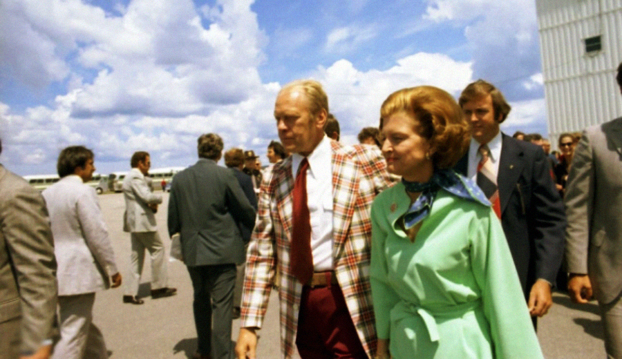 A man in a red plaid jacket and a woman in a light green dress with a blue scarf walk outside among a group of people under a partly cloudy sky.