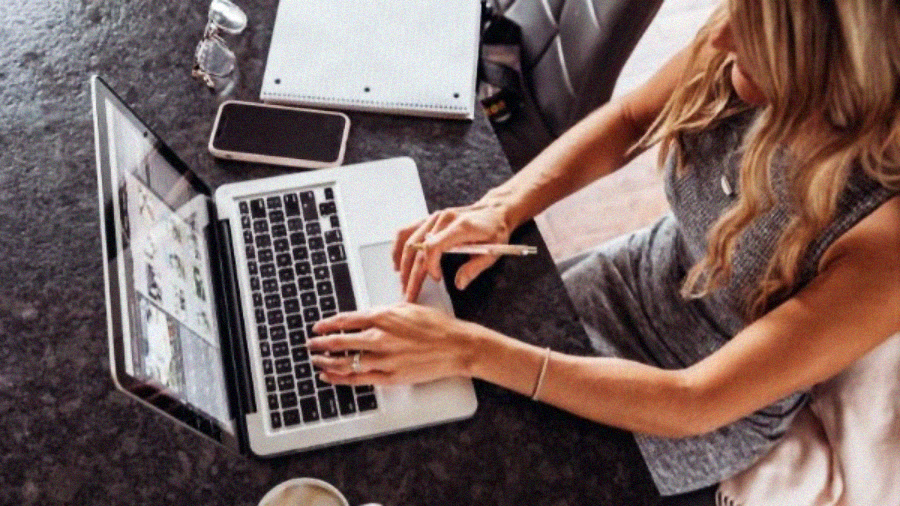 A woman sits at a table working on a laptop, using one hand to type and the other to hold a pen. A smartphone, notebook, and a cup of coffee are on the table beside her.