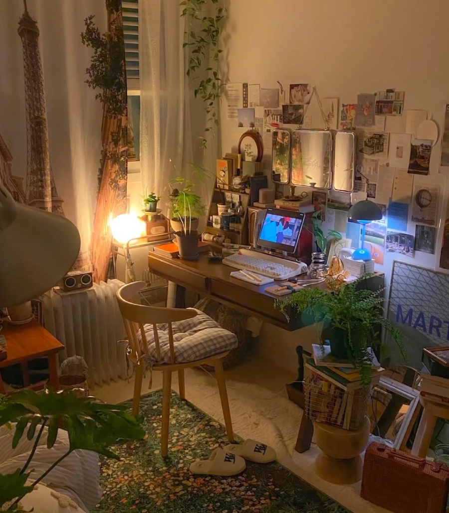A cozy, warmly-lit bedroom with a desk cluttered with plants, books, and a computer. There are wall decorations, mirrors, and postcards above the desk, and a chair with a cushion. A large plant and soft slippers are on a patterned rug.