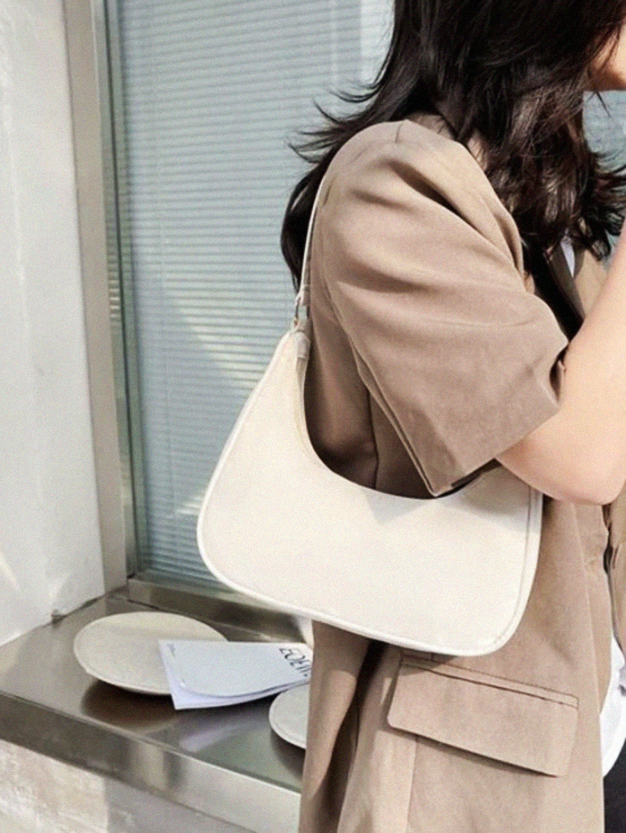 A woman with wavy dark hair, wearing a light brown blazer, stands by a window carrying a white shoulder bag. Sunlight casts shadows onto the wall and window behind her.