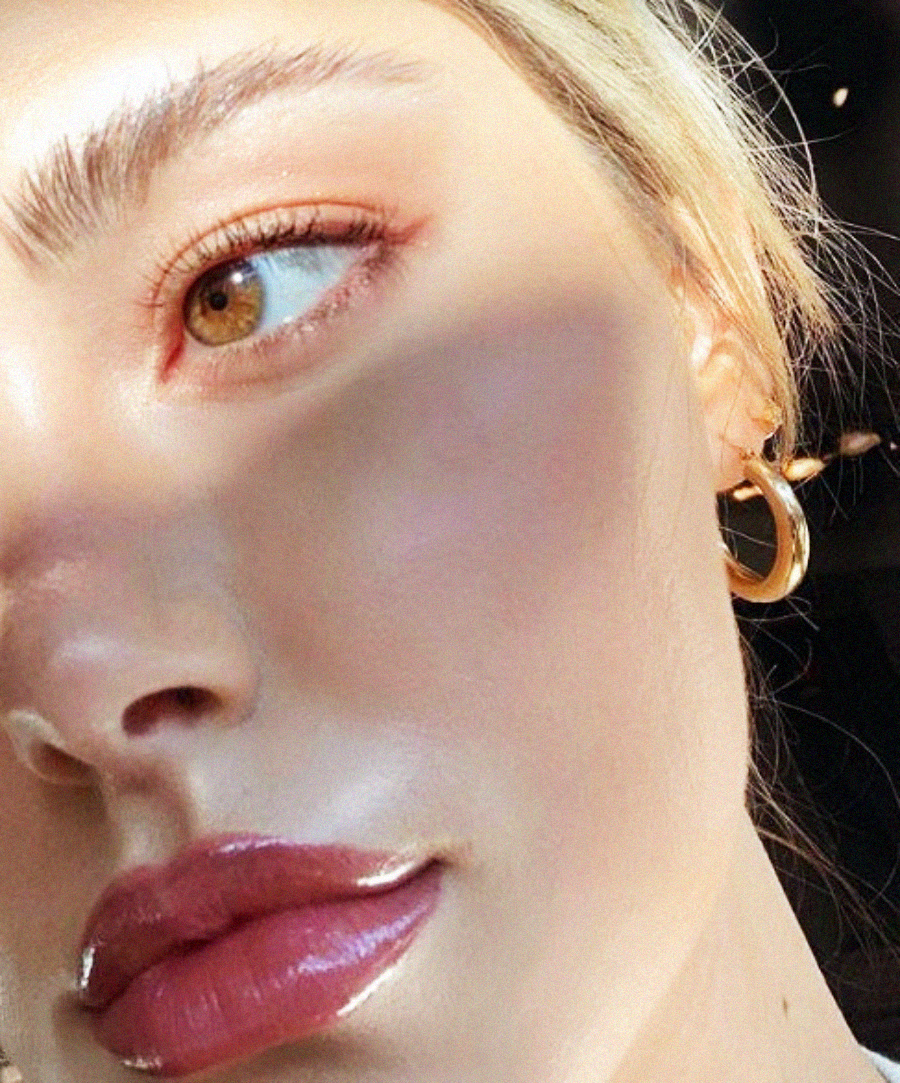 A close-up of a woman's face in sunlight, highlighting her glossy pink lips, brown eye with shimmery eyeshadow, golden earring, and smooth, radiant skin. Shadows partially cover her face.