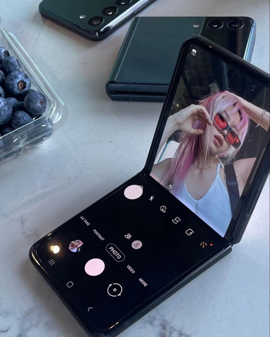 A foldable smartphone displays a camera app with a woman posing for a selfie. She has pink hair and red sunglasses. Another folded phone and a container of blueberries sit on a marble surface nearby.