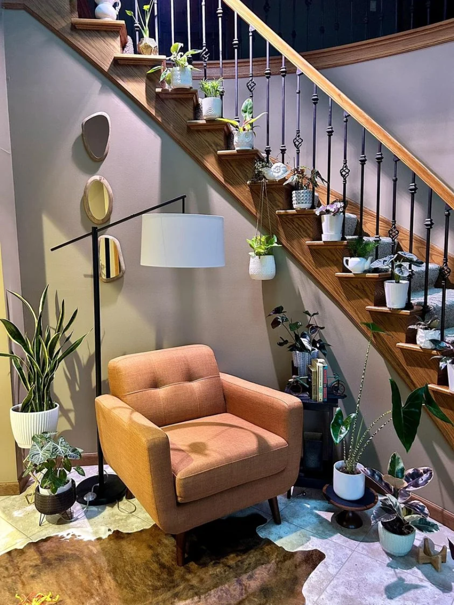 A cozy reading nook with a mid-century orange armchair, a modern floor lamp, and many potted plants arranged on the floor and along the wooden staircase, creating a lush, inviting atmosphere.