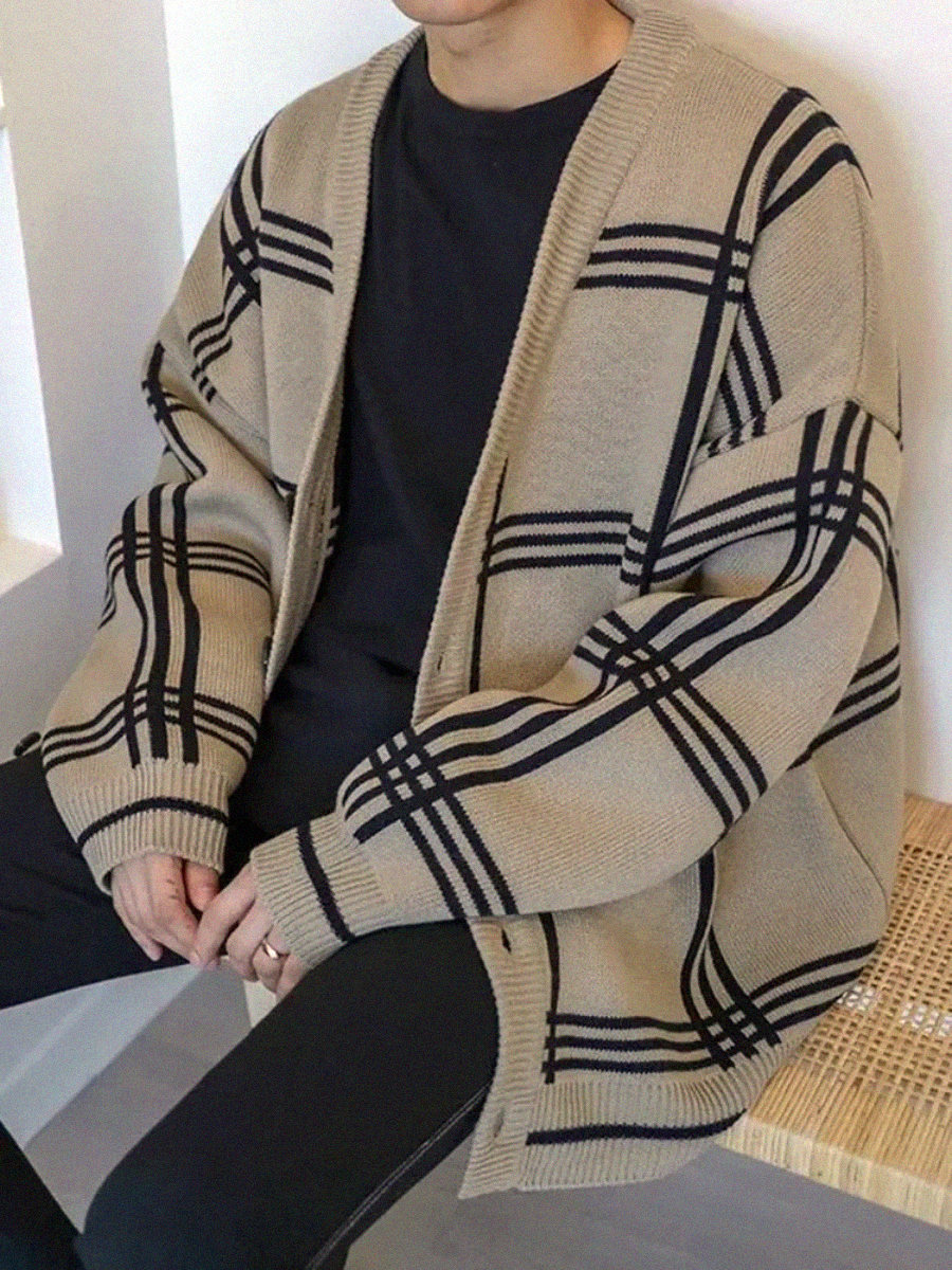 A person is seated wearing an oversized beige cardigan with black plaid stripes over a black shirt and black pants. The person's face is not visible.