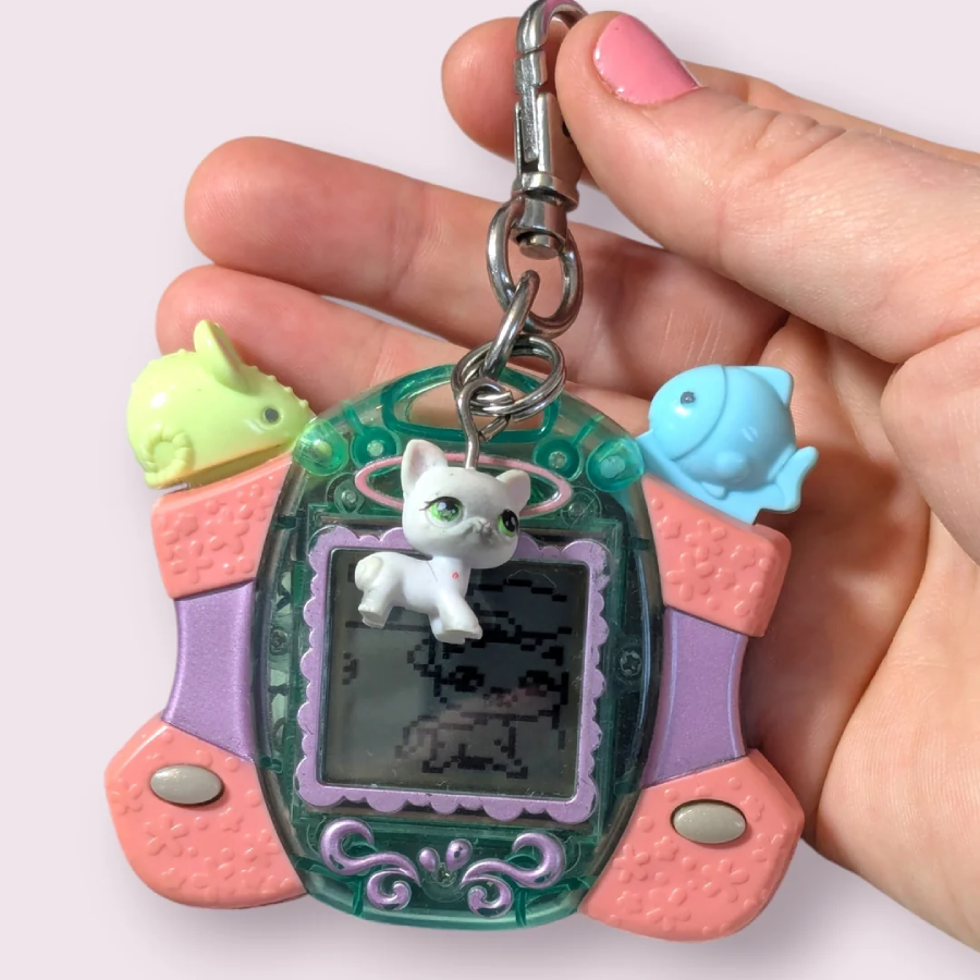 A hand holding a pastel keychain virtual pet toy with a cat charm and two fish charms attached. The person has pink nail polish on their thumbnail. The device has a screen showing a pixelated pet.