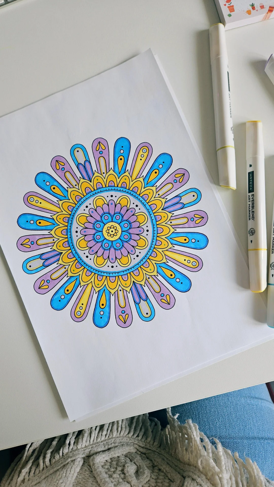 A colorful mandala drawing on white paper features shades of blue, yellow, and purple. It is surrounded by markers and placed on a white desk next to a person's leg covered with blue jeans and a fringed blanket.