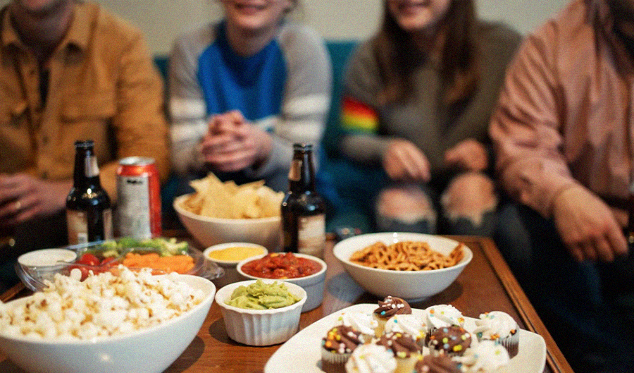 Four people sit together on a couch, smiling, with a wooden table in front of them filled with snacks like popcorn, chips, guacamole, salsa, cupcakes, pretzels, vegetables, and drinks. The image is brightly lit and casual.