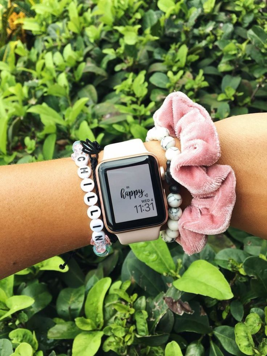 A wrist with a smartwatch displaying "happy" and the time 11:31, wearing a pink scrunchie, beaded white bracelets, and a letter bead bracelet spelling "CHILLIN," against a background of green plants.