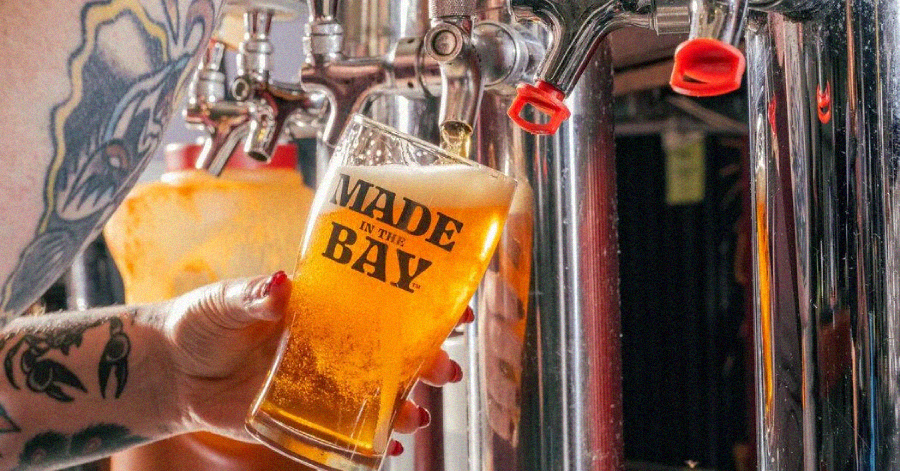 A tattooed person fills a glass of beer from a tap. The glass, held at an angle, features the words "Made in the Bay" and is topped with a foamy head. Bright lighting highlights the golden color of the beer.