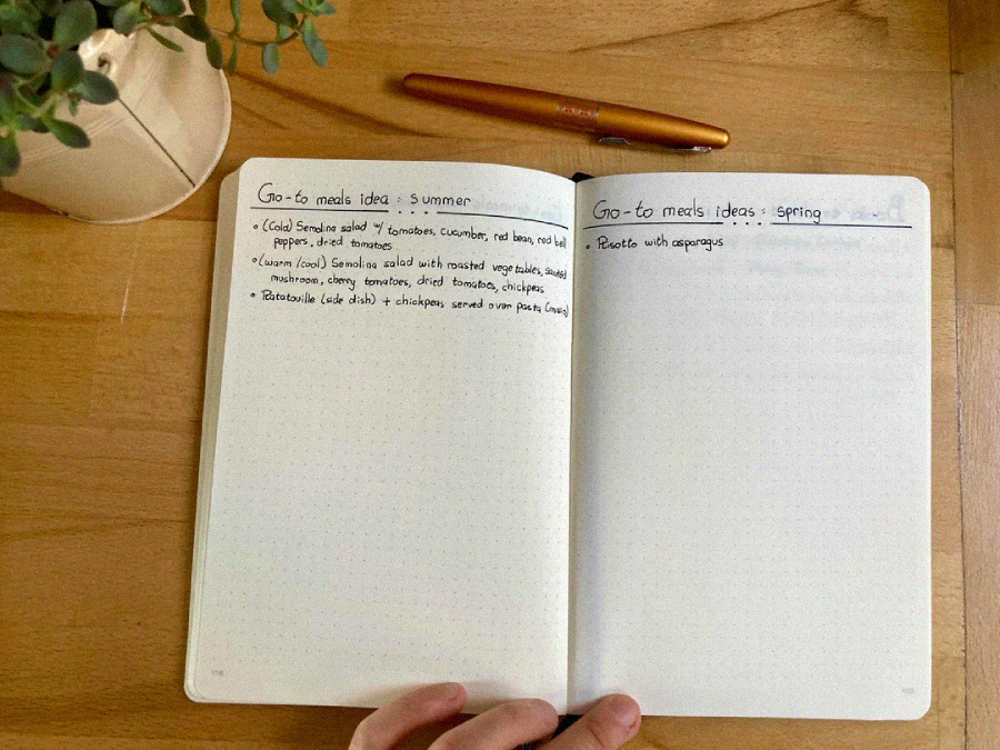 An open notebook on a wooden table shows handwritten meal ideas for summer and spring. A plant and a pen rest nearby. A hand holds the notebook at the bottom edge. The summer list includes salads and a chickpeas recipe.