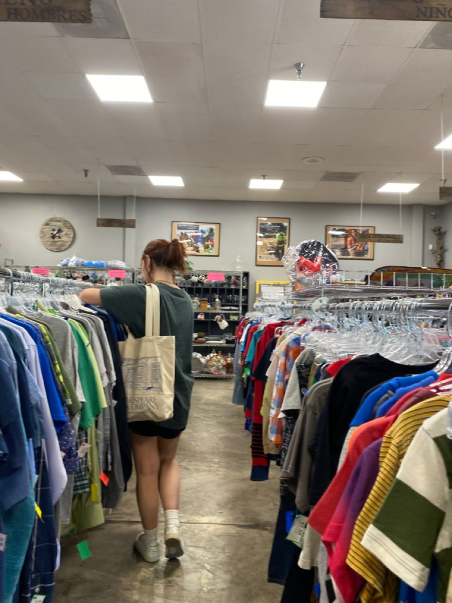 A person with a beige tote bag browses through racks of colorful clothes in a thrift store, with bright lighting and framed pictures on the walls.