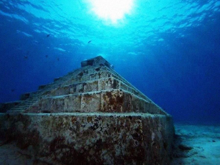 Underwater view of a large, ancient stone pyramid, partially covered in algae, with sunlight filtering through the water above and small fish swimming nearby.