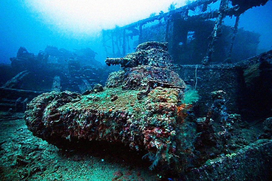 A rusted, coral-covered tank sits on the ocean floor amid the wreckage of a sunken ship, surrounded by blue water and marine life.