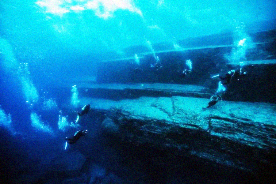 Several scuba divers swim near large, underwater stone formations that resemble steps or terraces, surrounded by deep blue water and rising bubbles.