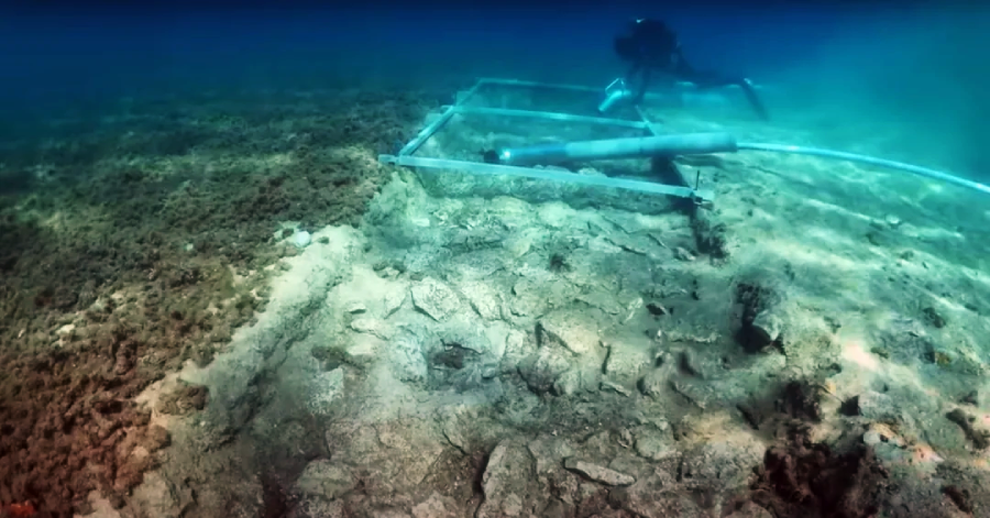 A scuba diver explores an underwater archaeological site, using a grid frame and excavation tools on a rocky seabed covered with scattered stones and patches of marine growth.