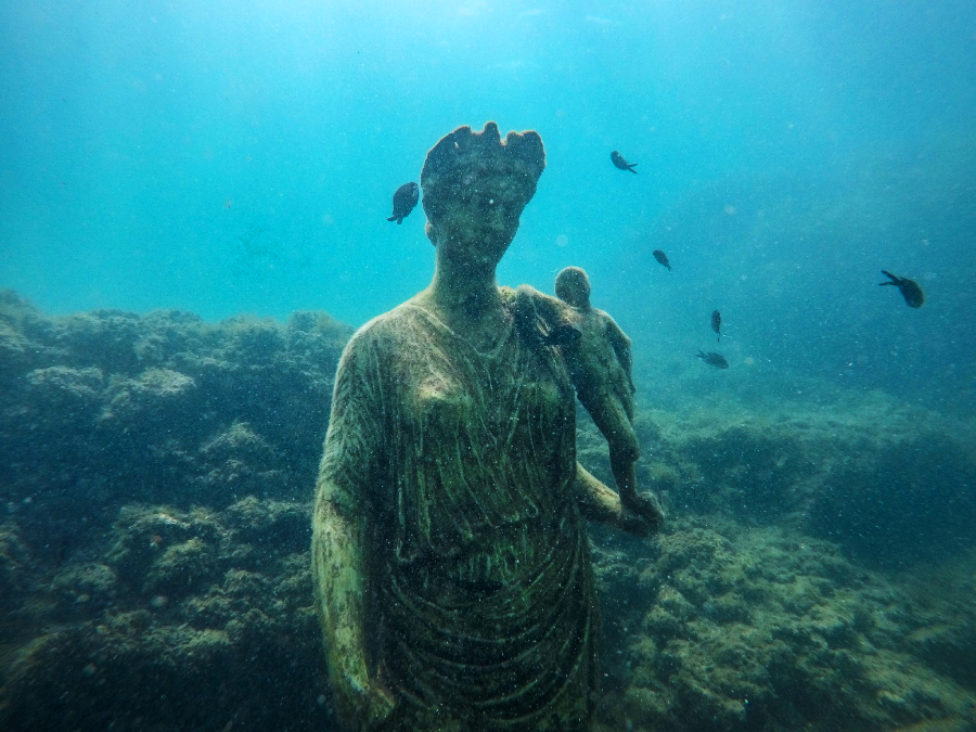 Underwater photo of a weathered statue of a woman holding a child, surrounded by small fish and marine life, with rocky seabed in the background and blue-green water.