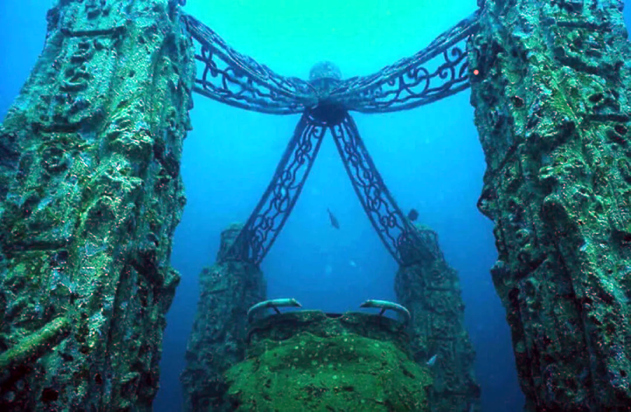Underwater view of a large, ornate metal structure covered in algae and marine growth, with tall pillars connected by curved, decorative beams, and blue ocean water in the background.