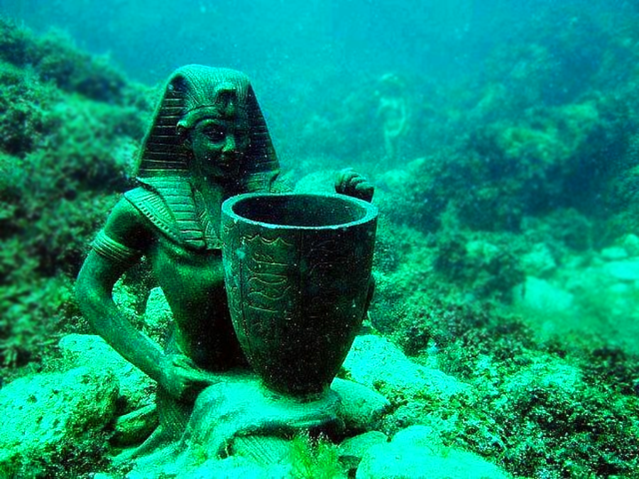A statue of an ancient Egyptian pharaoh kneeling and holding a large vessel sits underwater, surrounded by rocks and aquatic plants, with another statue faintly visible in the background.